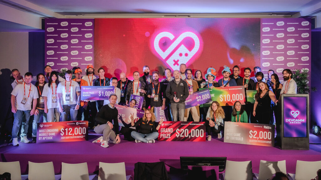 A photo from the DevGAMM Awards taken at the end of the show, with all the winners from the different categories standing on stage while holding their award checks. The DevGAMM Awards logo is shown on a screen behind the winners. In the picture, John and Jorge can be seen wearing their Whales And Games and Townseek-themed hats while holding the award check for the Best Game from Portugal.