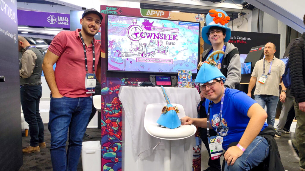 A photo of the team and José Castanheira standing by the Townseek and Exophobia booth at the GDC Expo, with a Townseek-themed hat on top of the chair, and the Wales booth behind in the background. The game booth features artwork of both games, with us standing on the side of our respective games.