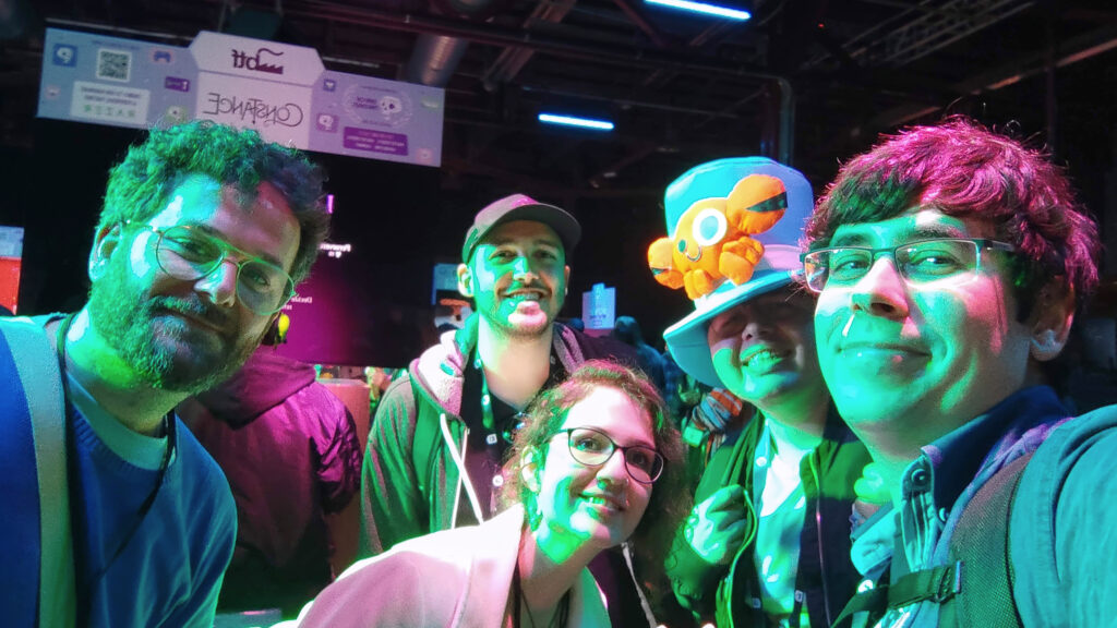 A photo of the team visiting the booth from the developers of Constance with Florian Köhne and Siba Gasser during the Day Of The Devs during GDC. Jorge and Moski are to the right of the picture, with Moski wearing his Whales And Games themed top hat. The photo is taken in a dark room, with purple and green lights illuminating the people in the photo.