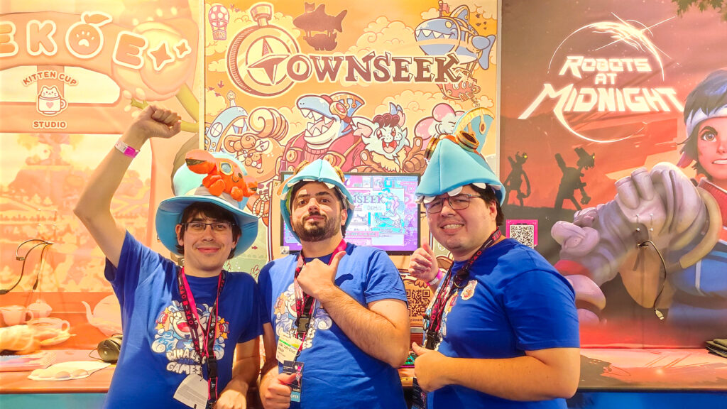 A photo of the team in front of the Townseek booth during gamescom. From left to right, Jorge, John, and Moski are posing in front of the Townseek stand while wearing blue shirts with the Whales And Games emblem on it. Jorge is wearing a Whales And Games themed top hat with a crab on it while doing a victory gesture with his arm, while John and Moski are wearing Townseek-themed hats and giving thumbs-up. The booth's artwork features the logo of the game, four of its characters, and the game's airship.