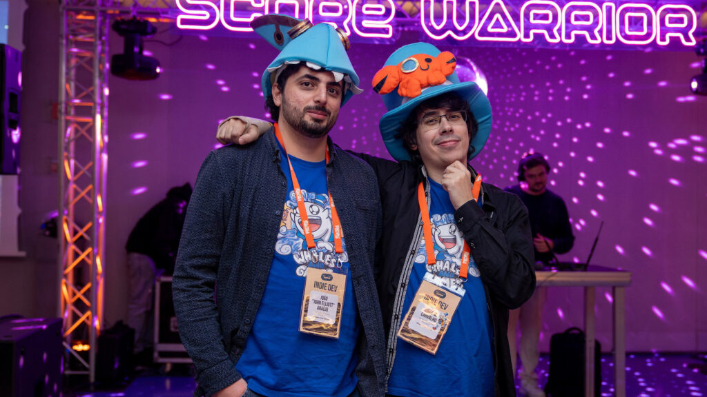 A photo from DevGAMM taken during the main party of the event. The photo features both John and Jorge, with Jorge with his arm wrapped around John while doing a thinking pose. Both are smiling at the camera while wearing their Whales And Games and Townseek-themed hats and their blue shirts with the Whales And Games emblem on them.