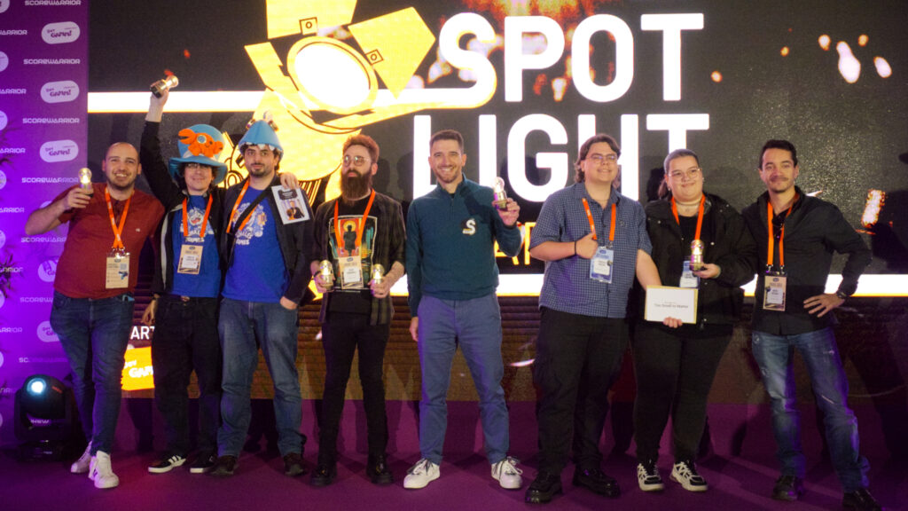 A photograph from the Spotlight Awards taken at the end of the show, with all the winners from the different categories standing on stage holding their trophy lightbulbs. The Spotlight Awards logo is shown on a screen behind us. In the picture, John and Jorge can be seen wearing their Whales And Games and Townseek-themed hats, while Jorge holds the trophy lightbulb in the air in a victory fist motion.